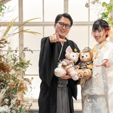 ドライフラワーに囲まれた温室風スタジオで、ぬいぐるみを抱えて笑う紋付袴の新郎と白無垢の花嫁。遊び心のある和装スタジオフォト