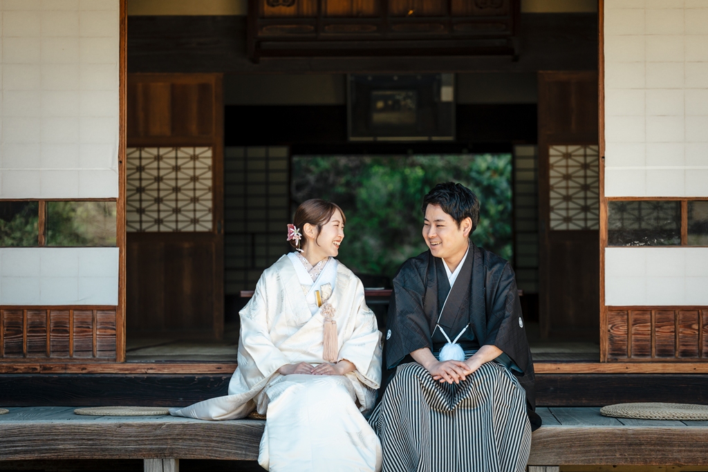 木造の縁側に腰掛けて笑い合う和装の新郎新婦