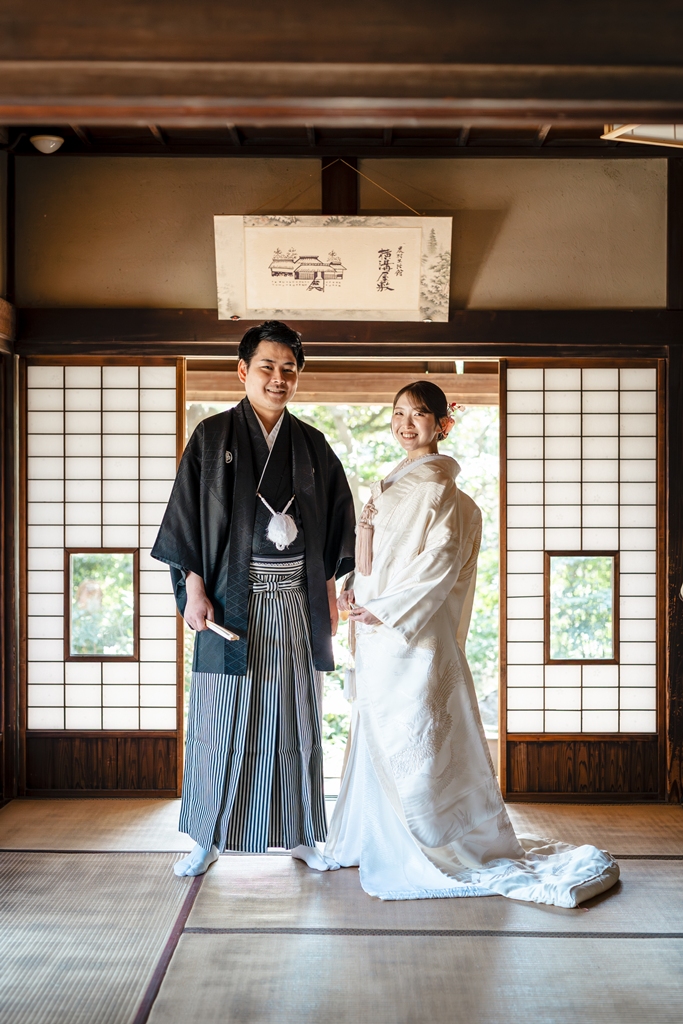 和室の入口で立ち姿を見せる白無垢と黒紋付袴の新郎新婦