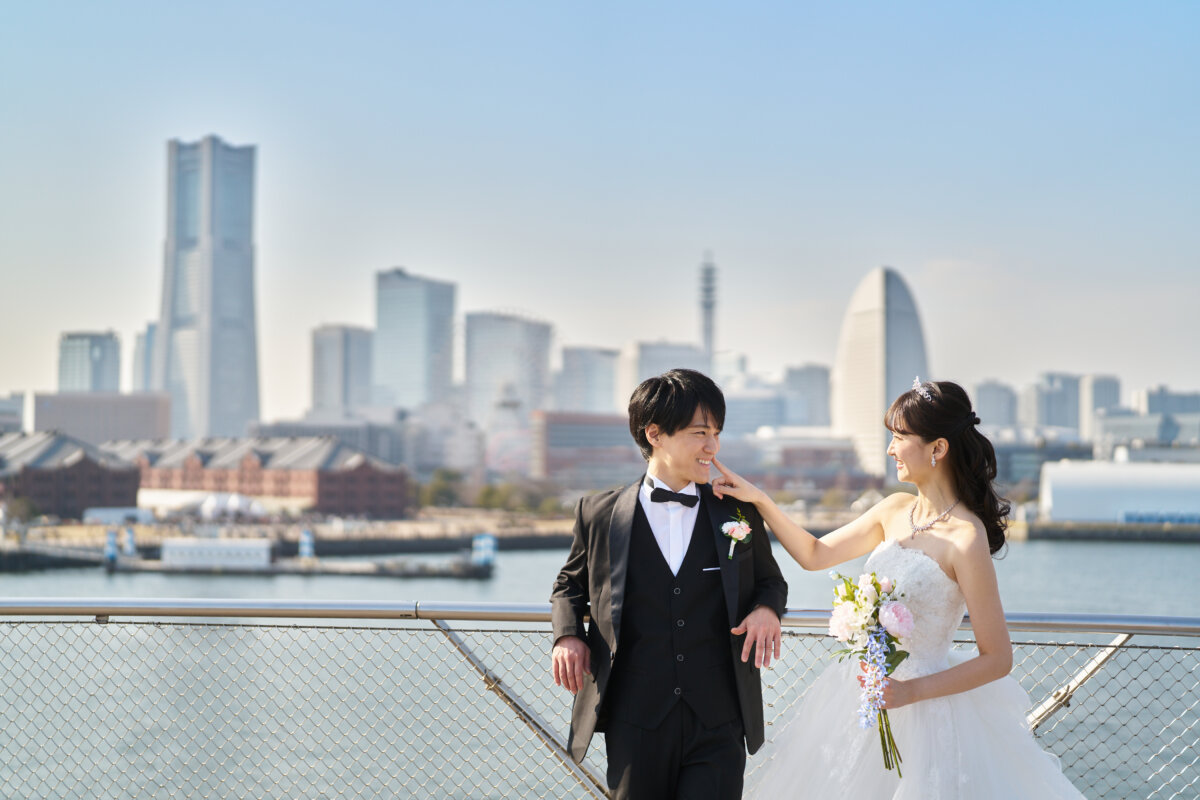 快晴の空とみなとみらいの高層ビル群を背景に立つタキシードとドレスの新郎新婦