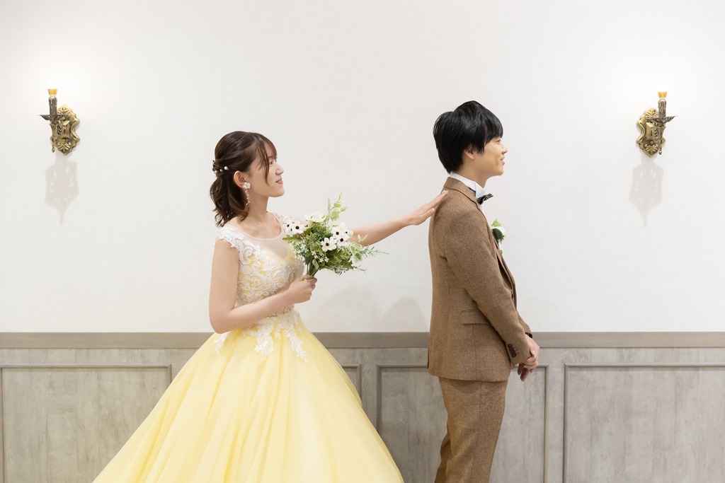 白い壁のスタジオでブラウンのスーツの新郎の肩をつつくブーケを持つ黄色いカラードレスの花嫁。横浜店のスタジオで撮るファーストミートの洋装前撮り