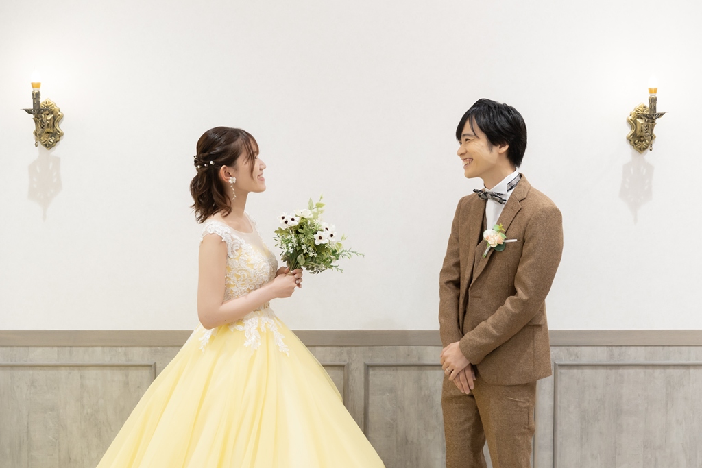 白い壁のスタジオで振り返って笑うブラウンのスーツの新郎とブーケを持つ黄色いカラードレスの花嫁。横浜店のスタジオで撮るファーストミートの洋装前撮り