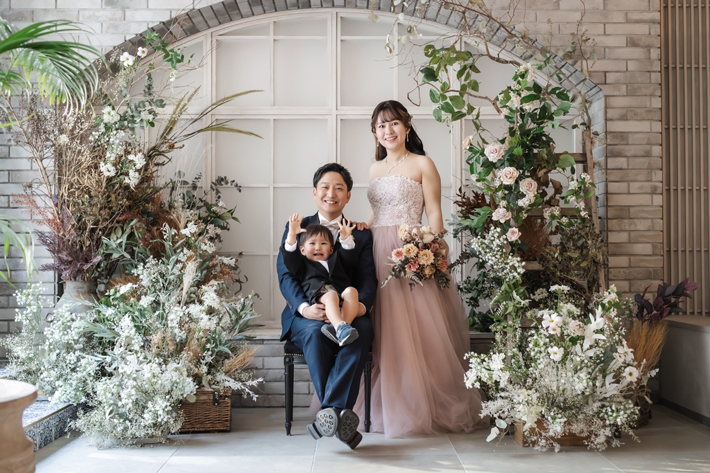 花で飾ったアーチ窓の前で幼い子どもを抱いて座る紺のスーツの新郎とピンクのカラードレスの花嫁。家族と一緒に撮る横浜店のスタジオフォト