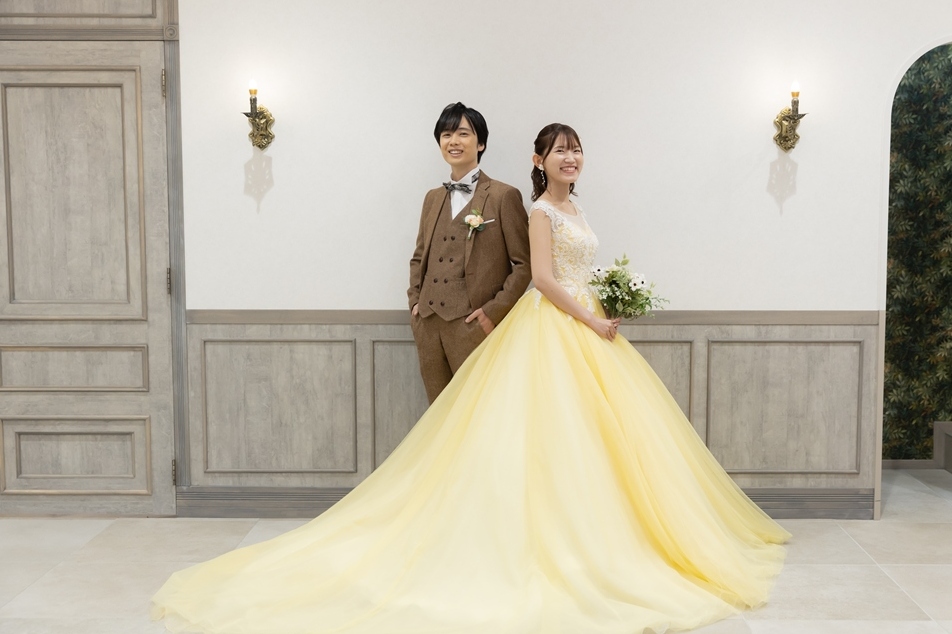 白い扉のスタジオで黄色いカラードレスのトレーンを広げて立つ花嫁とブラウンのスーツの新郎を引きでとらえた一枚。横浜店のスタジオで撮る洋装前撮り