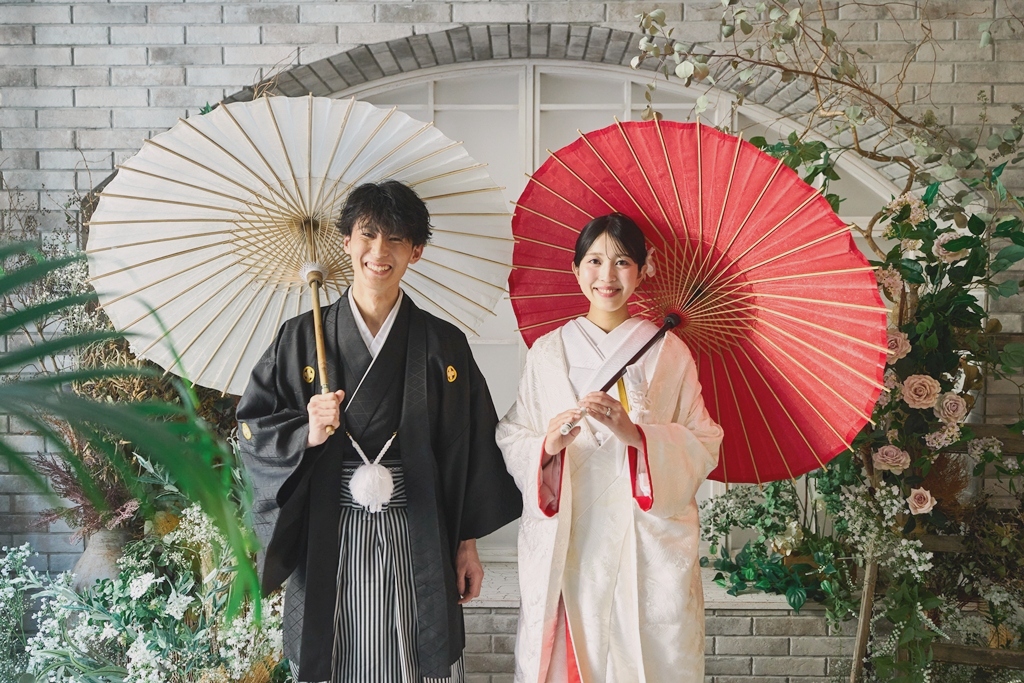 花で飾ったアーチ窓の前で白い和傘と赤い和傘を差す黒紋付袴の新郎と白無垢の花嫁。横浜店のスタジオで撮る和装前撮り