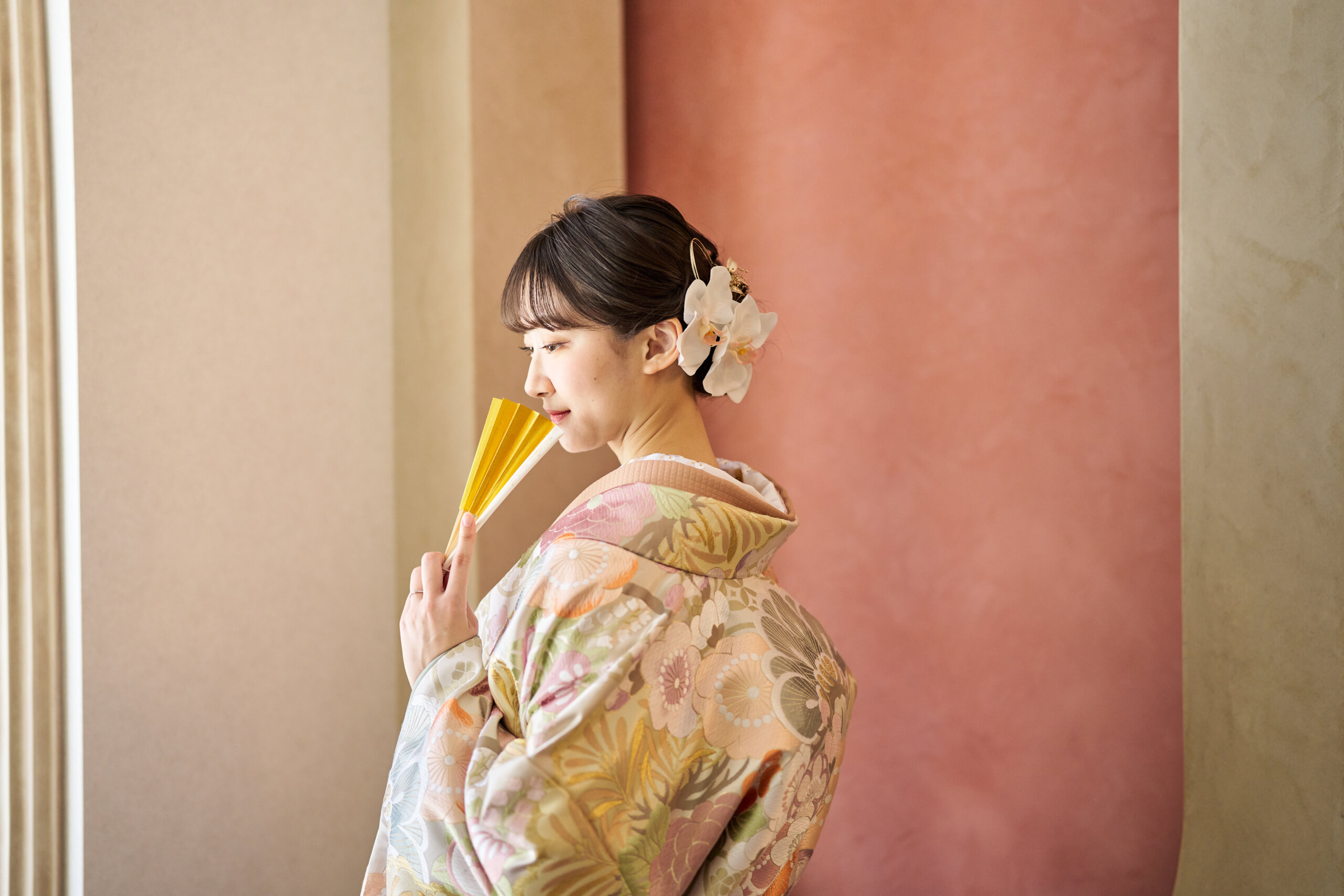 テラコッタ色の壁の前で金の扇子を持って横を向く花柄の色打掛の花嫁。横浜店のスタジオで撮る和装前撮りのブライダルポートレート