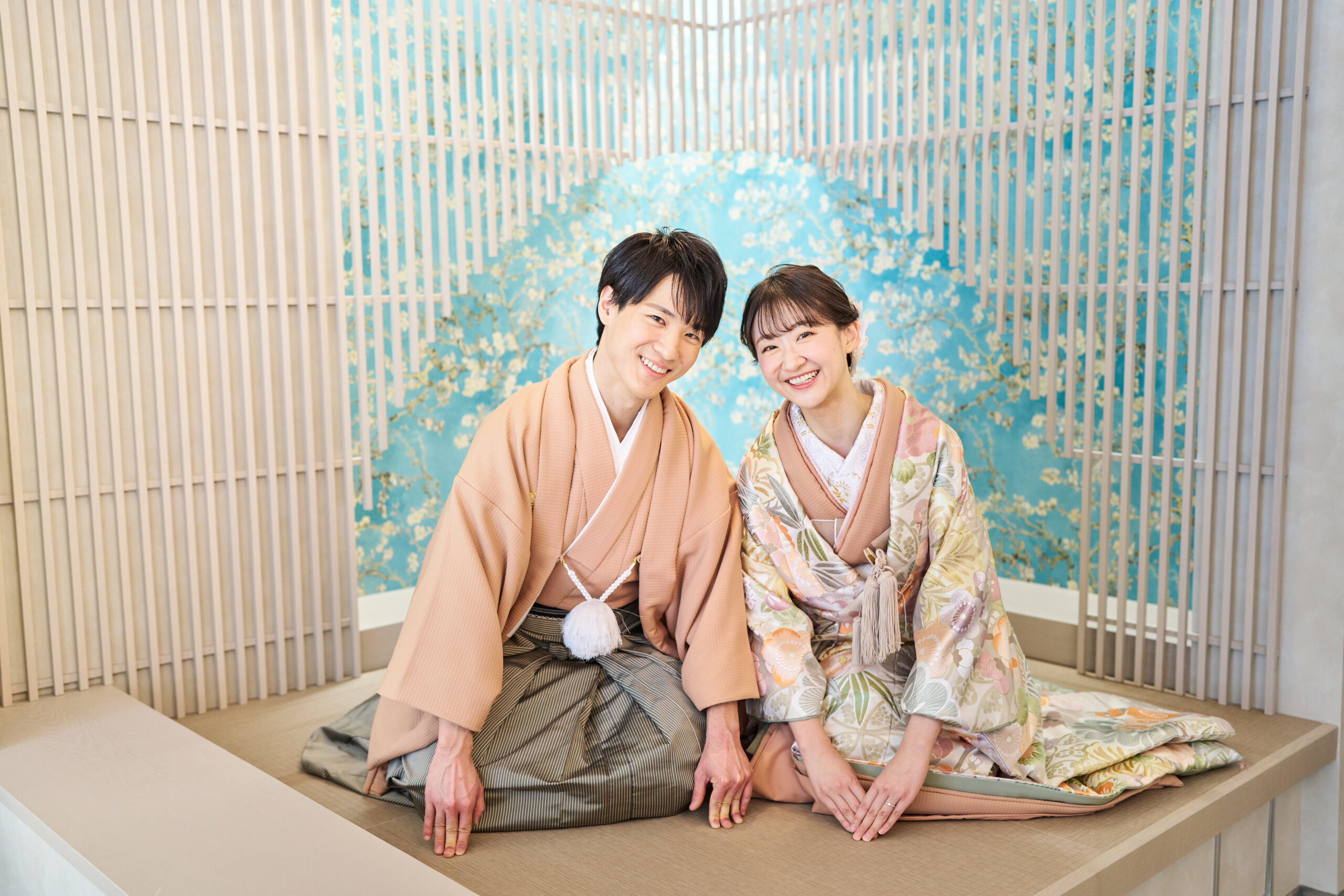 青い花柄のパネルの前に正座して笑うピンクベージュの羽織袴の新郎と花柄の色打掛の花嫁。横浜店のスタジオで撮る和装前撮り