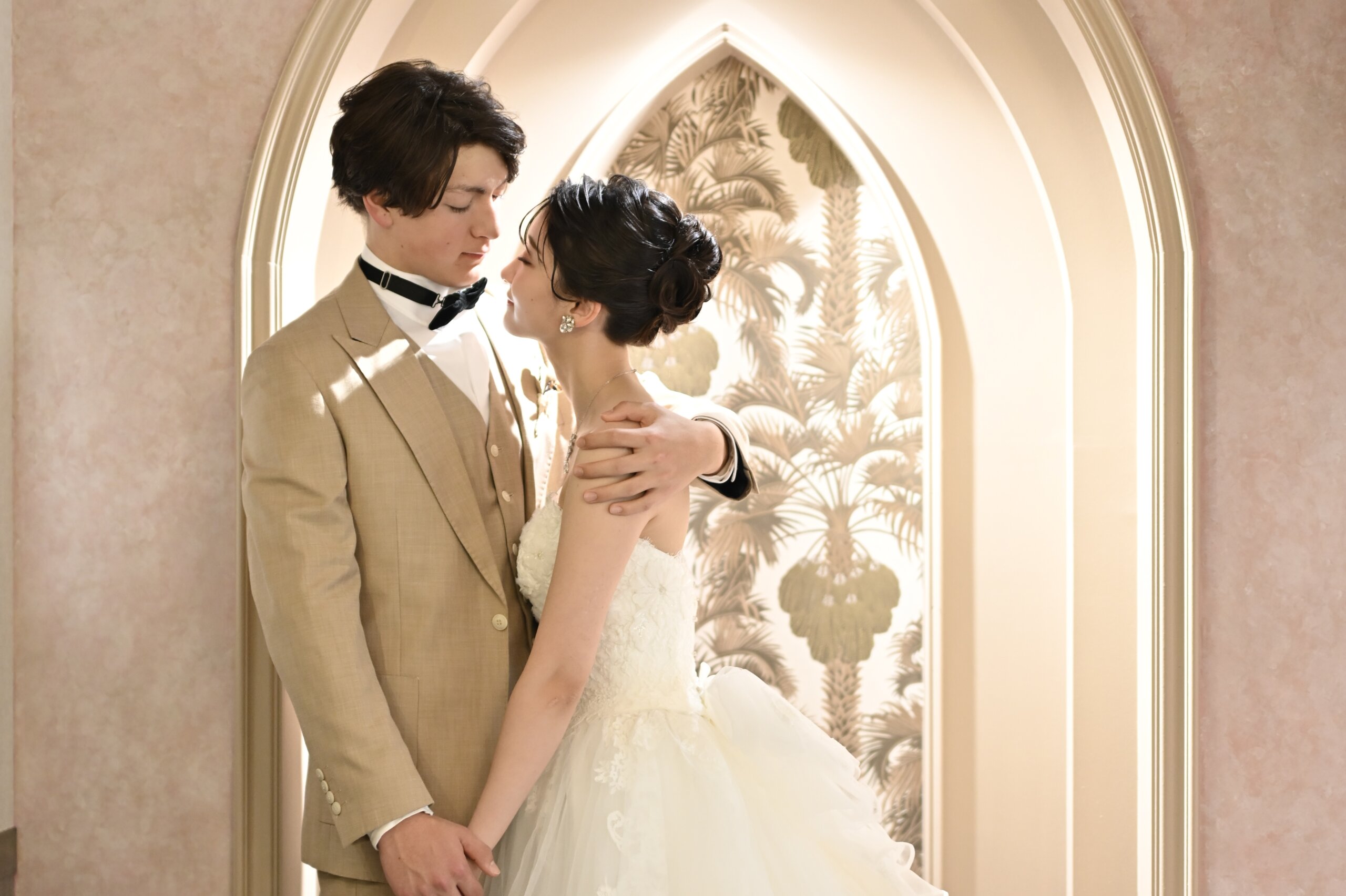 ヤシの葉の壁紙のアーチの前で額を寄せ合うベージュのスーツの新郎とウェディングドレスの花嫁。横浜店のスタジオで撮る洋装前撮り
