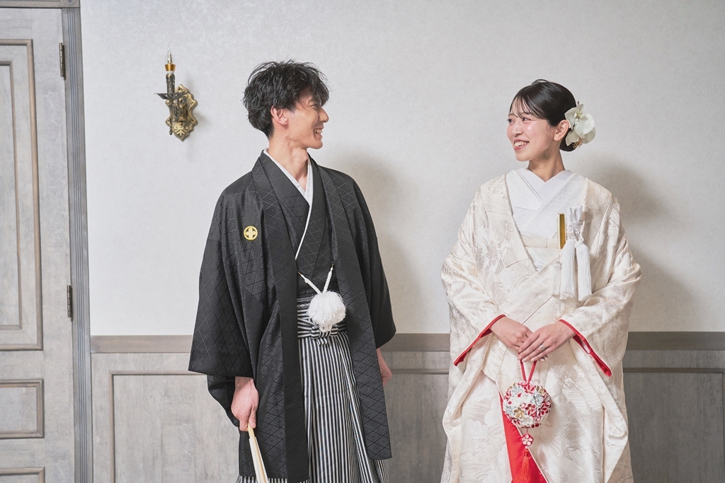 白い壁の前で向かい合って笑う黒紋付袴の新郎と白い色打掛の花嫁。横浜店のスタジオで撮る和装のスタジオフォト