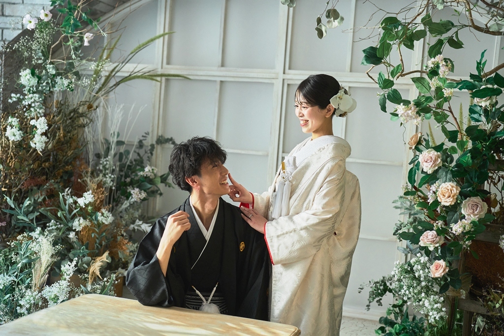 花で飾ったアーチ窓の前でテーブルに座る新郎の頬に手を添える白い色打掛の花嫁。横浜店のスタジオで撮る和装前撮り