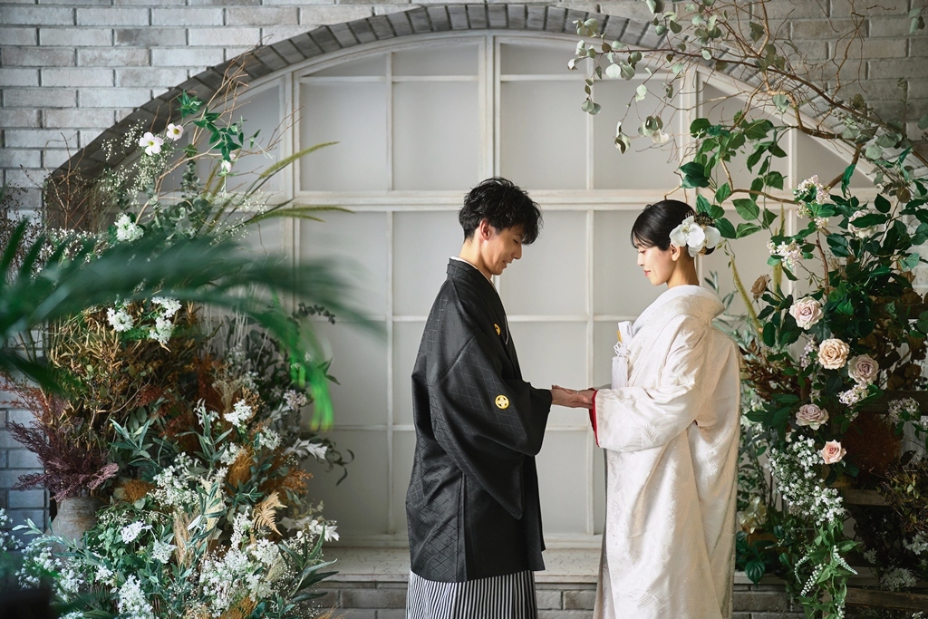 花で飾ったアーチ窓の前で手を取り合う黒紋付袴の新郎と白い色打掛の花嫁。横浜店のスタジオで撮る和装のスタジオフォト