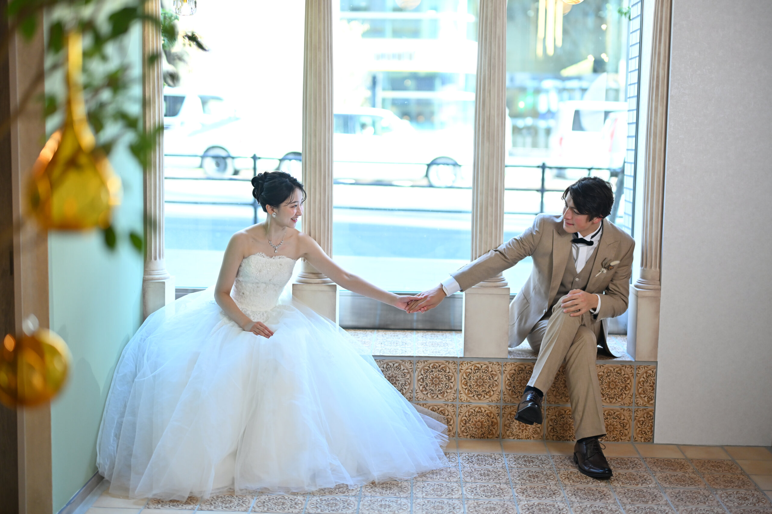 アーチ窓の窓辺に座って手をつなぐウェディングドレスの花嫁とベージュのスーツの新郎。横浜店のスタジオで撮る洋装のスタジオフォト