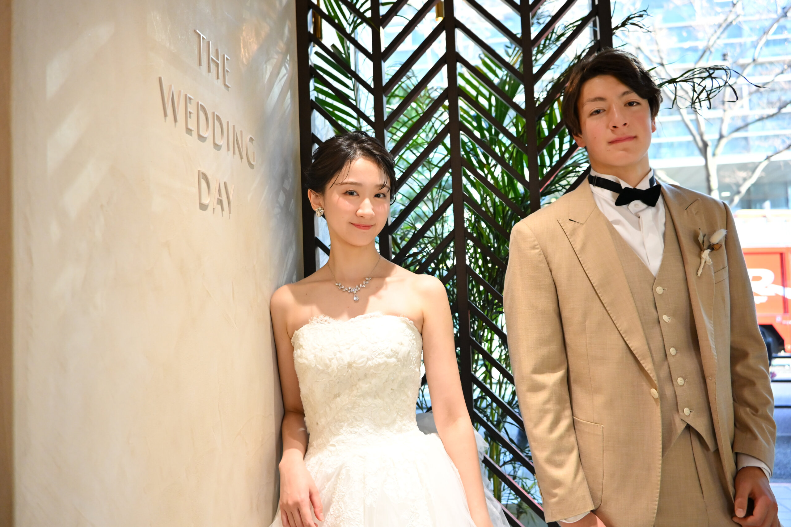 『THE WEDDING DAY』の文字の壁の前で微笑むベージュのスーツの新郎とウェディングドレスの花嫁。横浜店のスタジオで撮る洋装前撮り