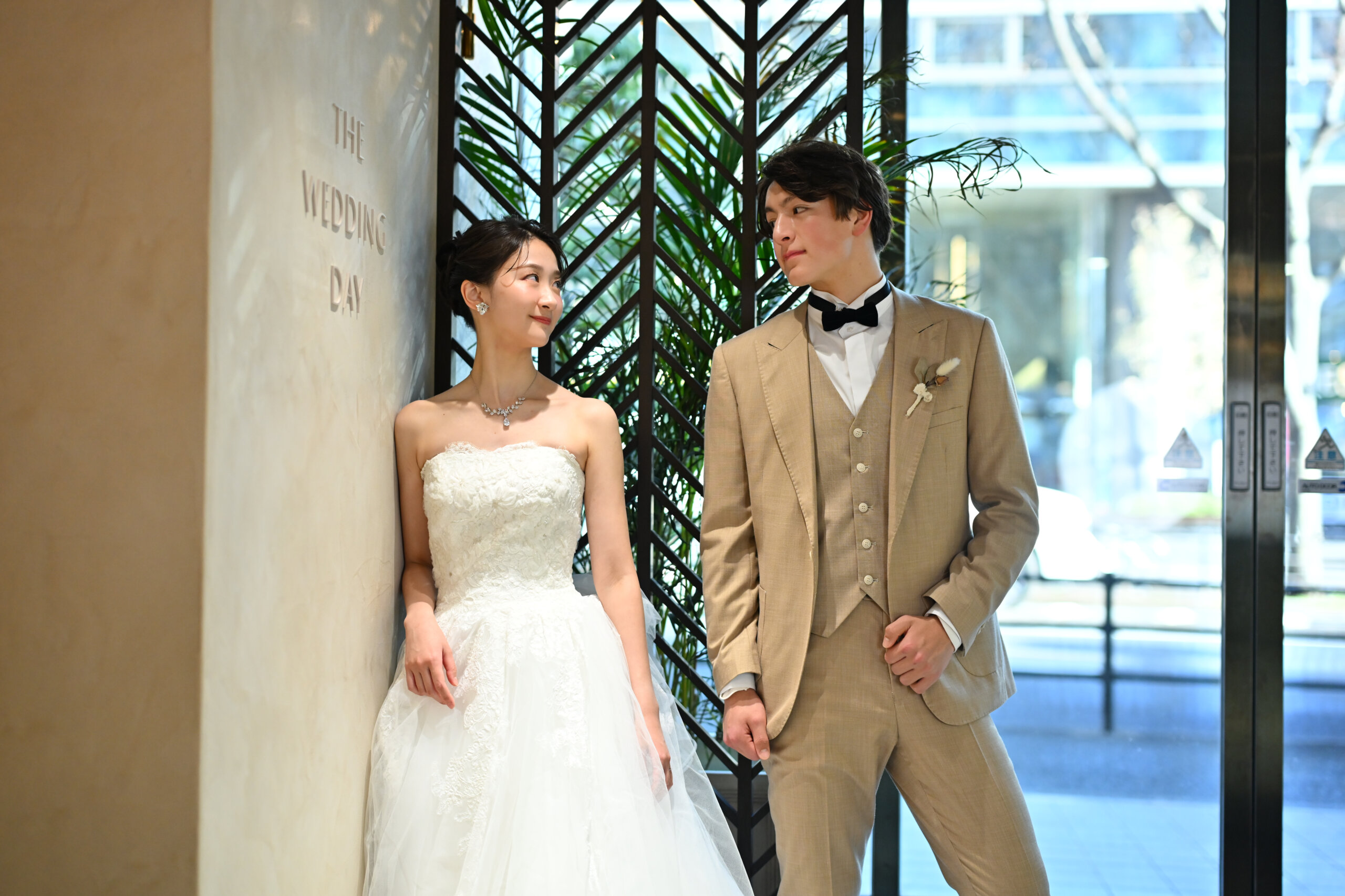 ヘリンボーンの格子の前で見つめ合うベージュのスーツの新郎とウェディングドレスの花嫁。横浜店のスタジオで撮る洋装のスタジオフォト