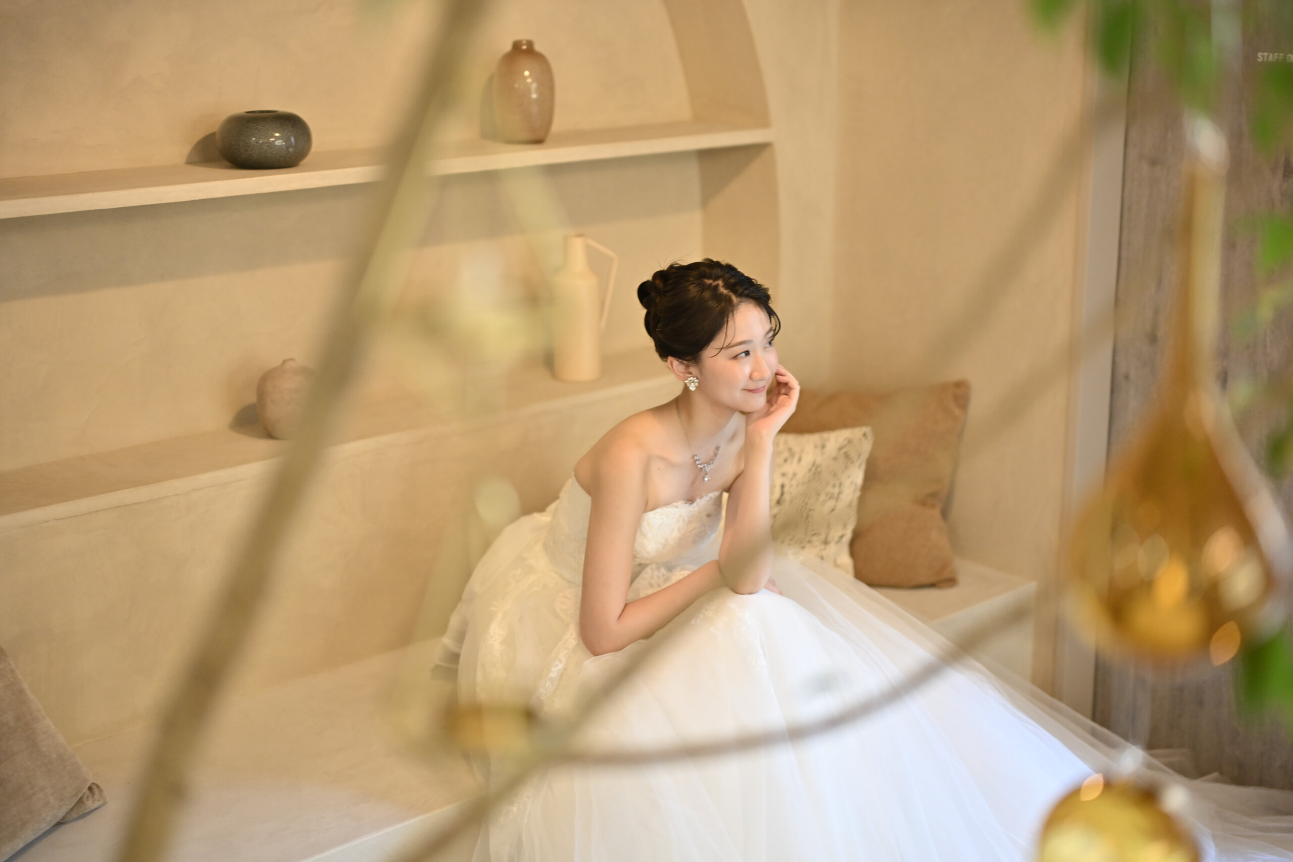 アーチ型の飾り棚のあるベージュの部屋のソファで頬杖をつくウェディングドレスの花嫁を緑越しにとらえた一枚。横浜店のスタジオで撮る洋装前撮りのブライダルポートレート