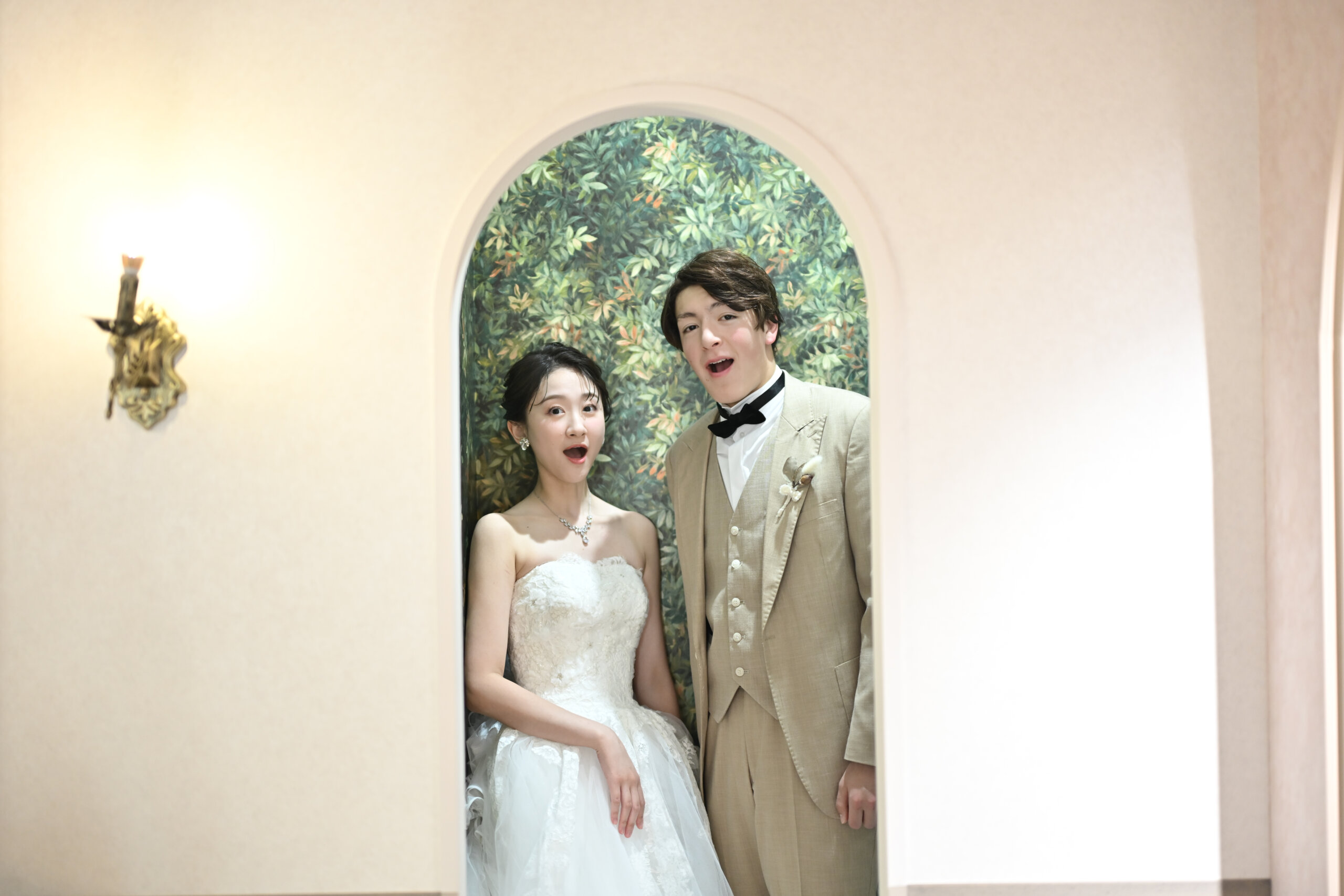 緑の壁紙のアーチの中で驚いた顔でおどけるベージュのスーツの新郎とウェディングドレスの花嫁。横浜店のスタジオで撮る洋装前撮りの遊び心のあるカット