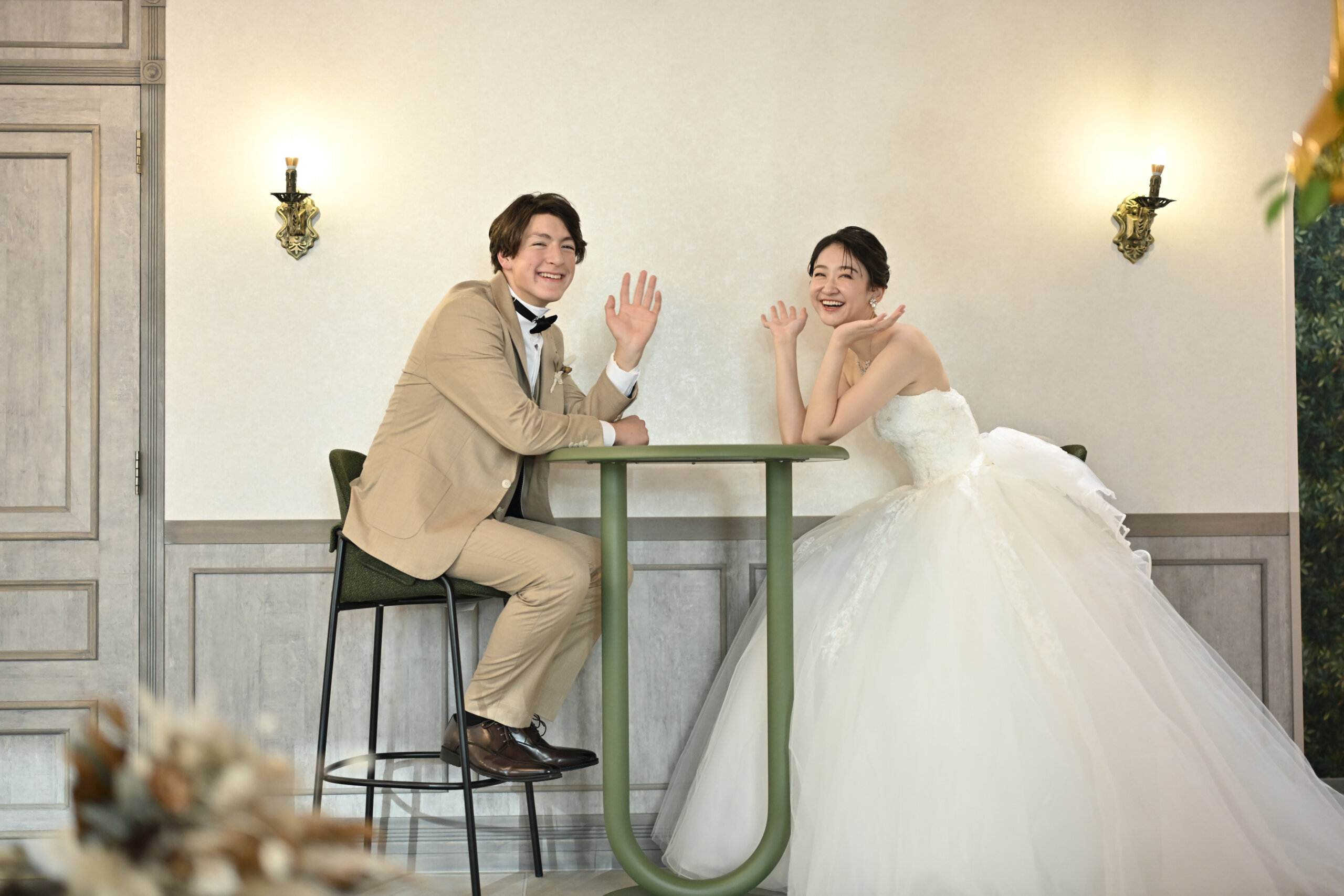 緑のテーブルで手を振っておどけるベージュのスーツの新郎とウェディングドレスの花嫁。横浜店のスタジオで撮る洋装前撮りの遊び心のあるカット