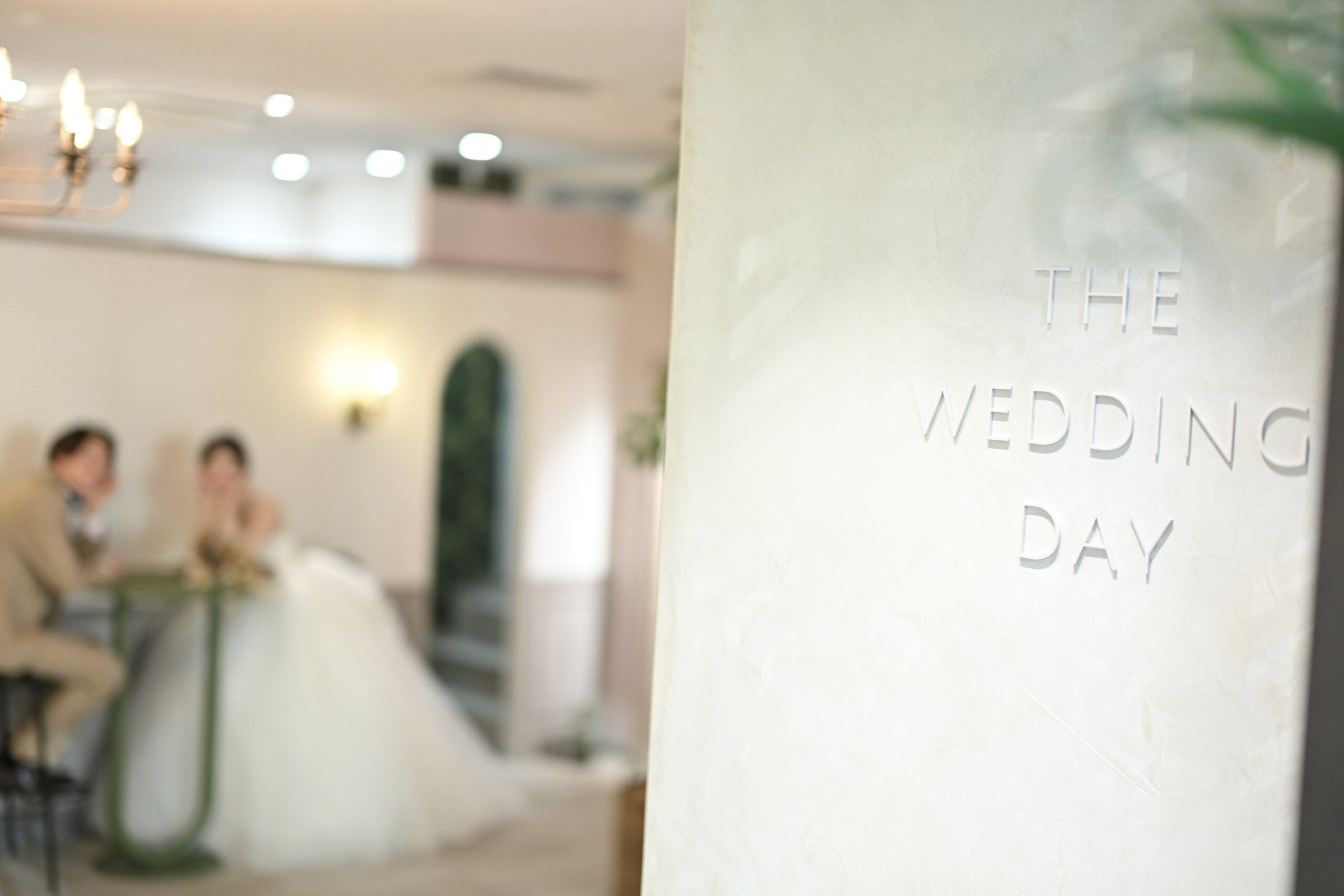 『THE WEDDING DAY』の文字の壁越しにテーブルで向かい合う新郎とウェディングドレスの花嫁をぼかしてとらえた一枚。横浜店のスタジオで撮る洋装前撮り