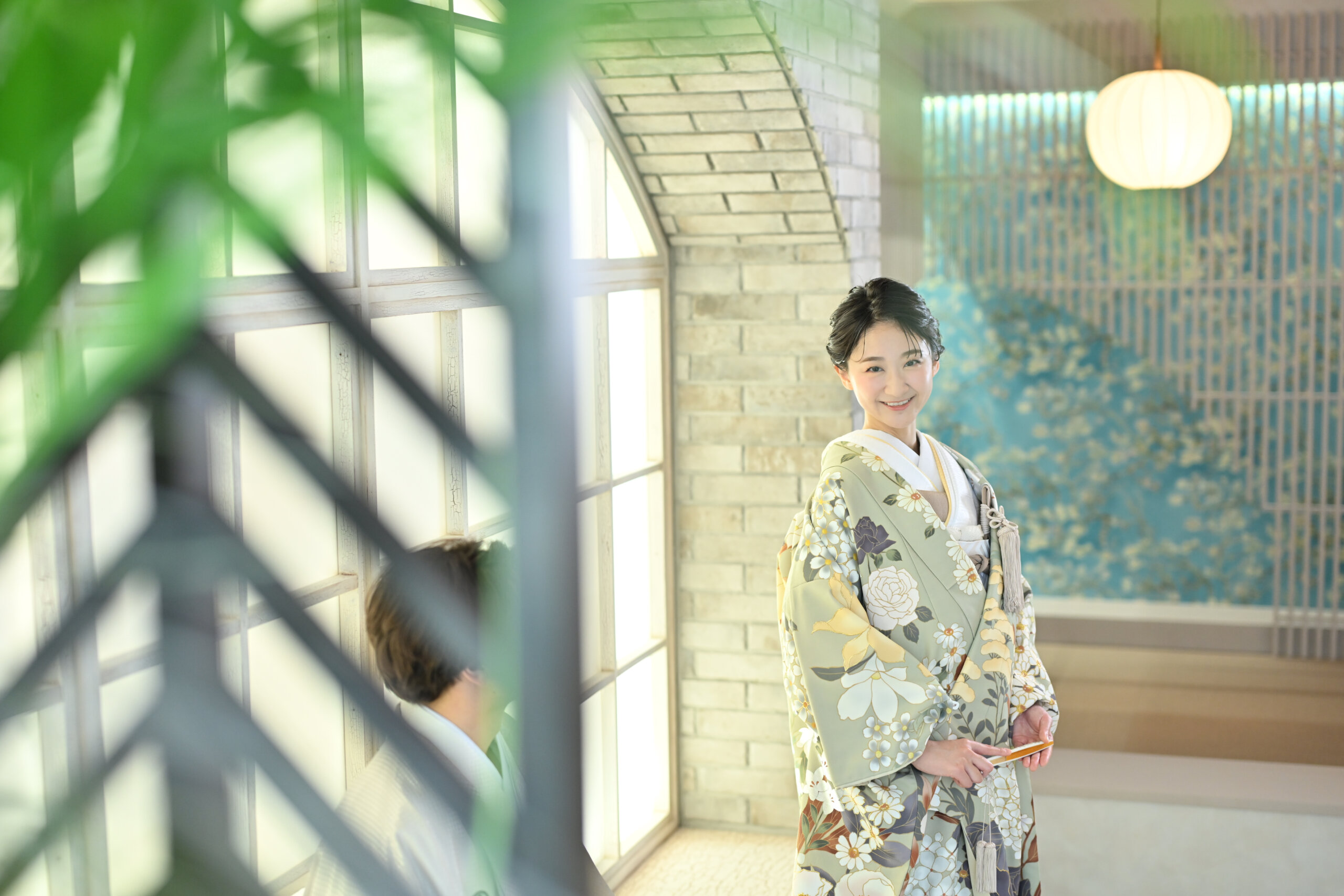 青い花柄のパネルの前で微笑む花柄の色打掛の花嫁を緑越しにとらえた一枚。横浜店のスタジオで撮る和装前撮りのブライダルポートレート