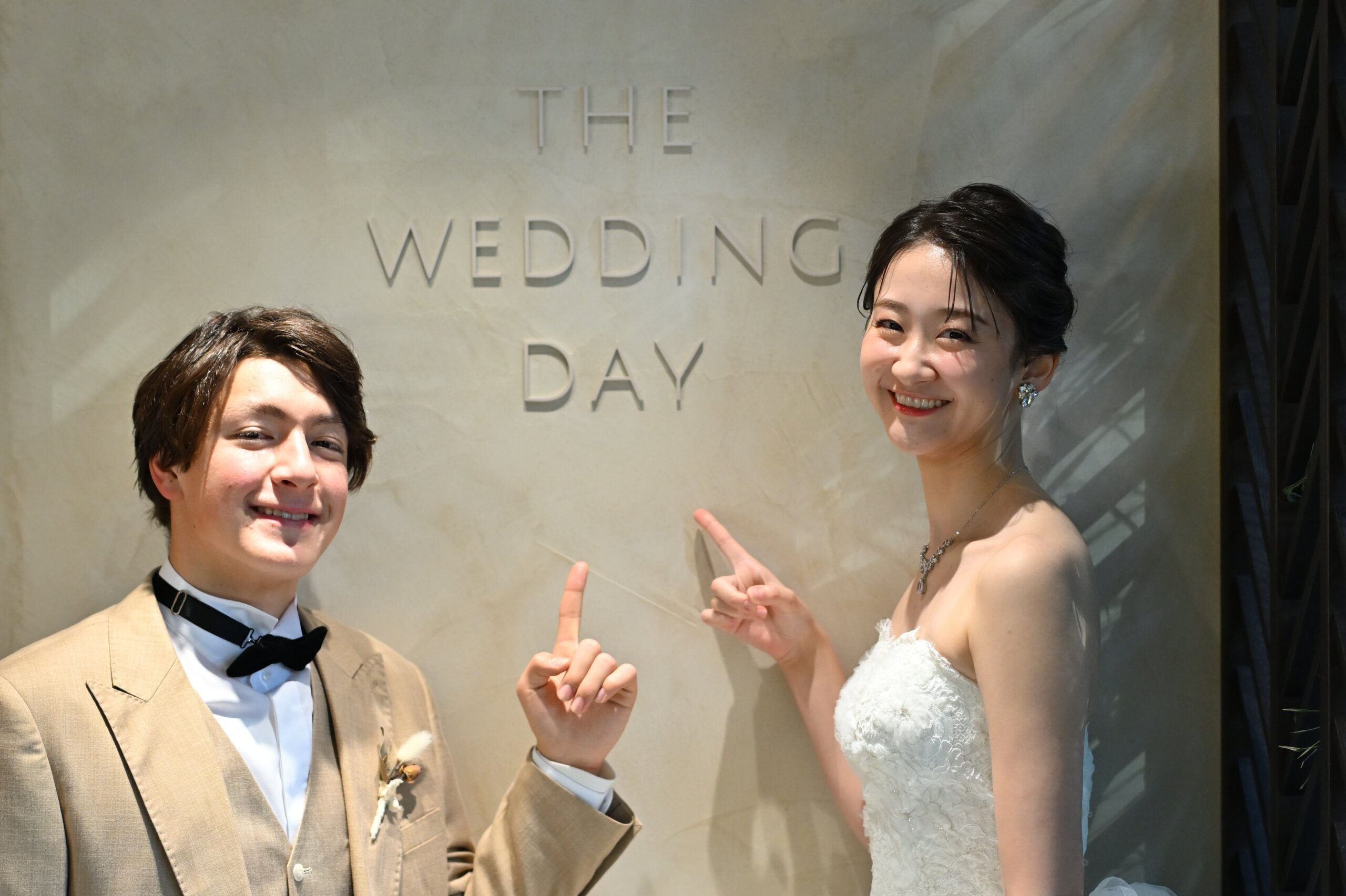 『THE WEDDING DAY』の文字を指差して笑うベージュのスーツの新郎とウェディングドレスの花嫁。横浜店のスタジオで撮る洋装前撮りの遊び心のあるカット