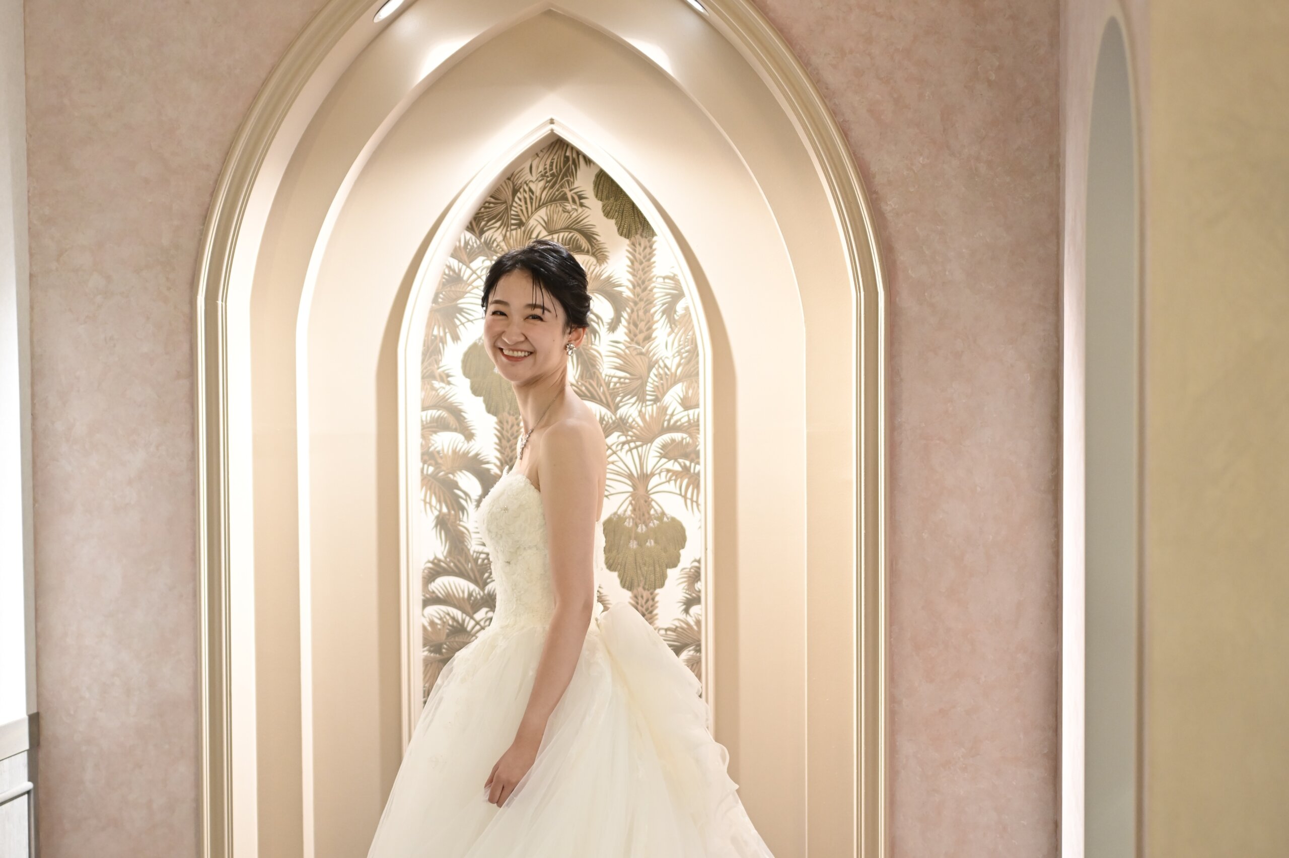 ヤシの葉の壁紙のアーチの前に立つウェディングドレスの花嫁を引きでとらえた一枚。横浜店のスタジオで撮る洋装前撮りのブライダルポートレート