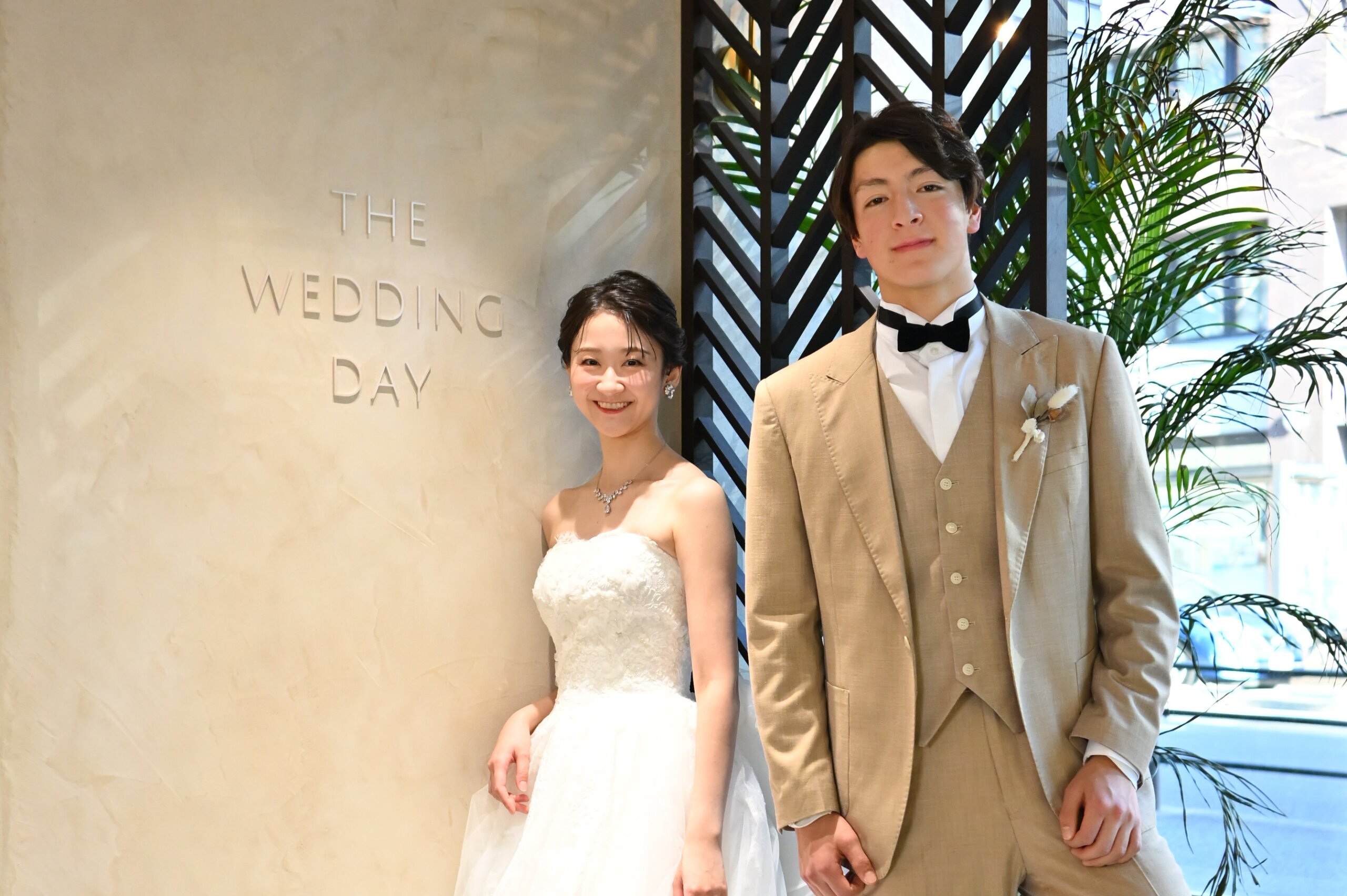 『THE WEDDING DAY』の文字の壁の前に並ぶベージュのスーツの新郎とウェディングドレスの花嫁。横浜店のスタジオで撮る洋装前撮り