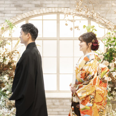 白レンガのアーチ窓とグリーンの装飾の前で向かい合う黒紋付袴の新郎とオレンジの色打掛の花嫁。横浜のスタジオで撮る和装前撮り
