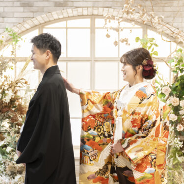 逆光のアーチ窓の前で、新郎が色打掛の花嫁の肩に手を添えて微笑み合うカット。スタジオの和装フォトウェディング