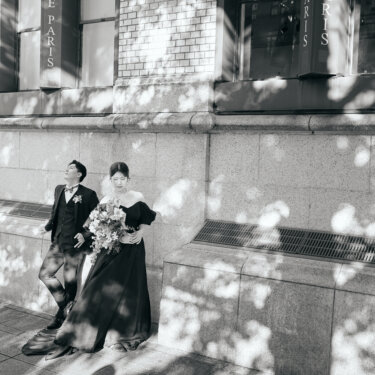 石造りの建物の壁にもたれて寄り添う新郎新婦。木漏れ日の影をモノクロで写した、レトロな雰囲気の結婚写真