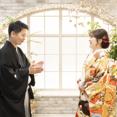 アーチ窓の前で手を合わせて笑い合う黒紋付袴の新郎とオレンジの色打掛の花嫁。スタジオで撮る和装前撮りの自然な一枚