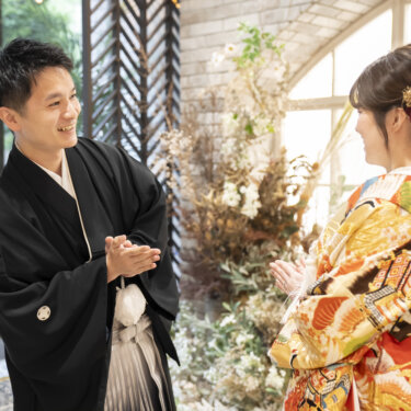 グリーンとドライフラワーを飾ったスタジオで、拍手をしながら向かい合う黒紋付袴の新郎と色打掛の花嫁。和装スタジオフォトの自然な笑顔