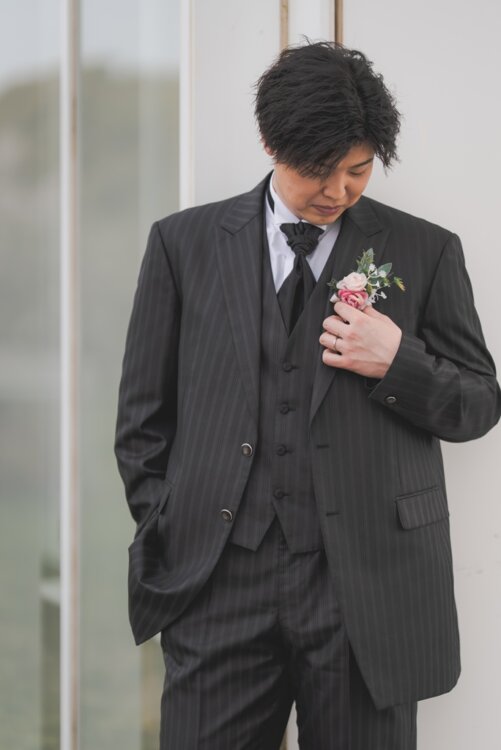 ストライプのブラックタキシードの胸元にピンクのブートニアを添えて、うつむき加減にほほえむ新郎。前撮りで映える大きなサイズのタキシード（ブラックラージ／衣装番号TP08）