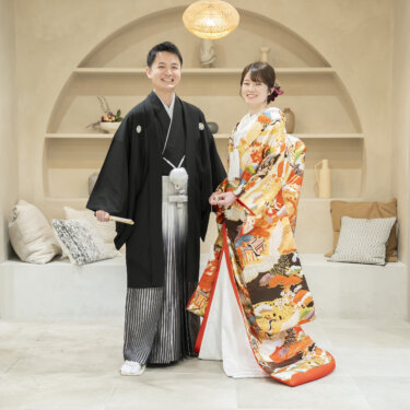 アーチ型のニッチとクッションのあるナチュラルなスタジオで並ぶ黒紋付袴の新郎とオレンジの色打掛の花嫁。和装スタジオフォトの全身カット