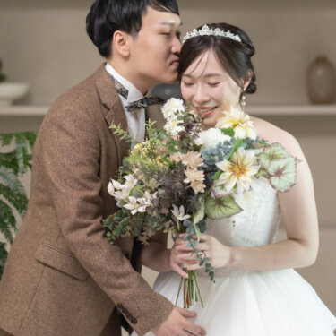 大きなブーケを抱えた花嫁の頬に新郎がキスをする瞬間。ティアラとウェディングドレスで撮る幸せな結婚写真