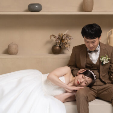 ベージュのソファで新郎のひざにもたれて眠るように目を閉じる花嫁。ウェディングドレスで撮るリラックスした寝そべりポーズのスタジオフォト