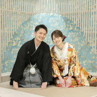 水色の花柄の壁と白い格子の前で正座して微笑む黒紋付袴の新郎と色打掛の花嫁。横浜のスタジオで撮る和装結婚写真