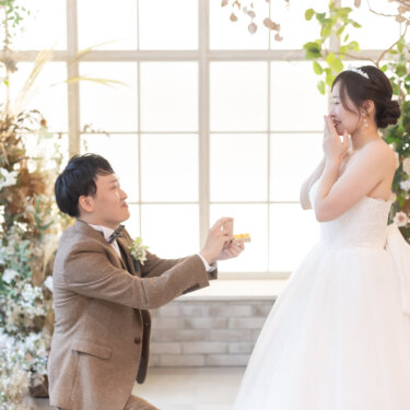 大きな窓とドライフラワーのセットで片膝をつき指輪を差し出す新郎と、口元を押さえて驚く花嫁。プロポーズを再現する結婚写真