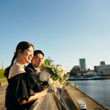 横浜港を背に、手すりにもたれて笑顔を見せる黒いドレスの花嫁とタキシードの新郎。大きなブーケが華やかな前撮り