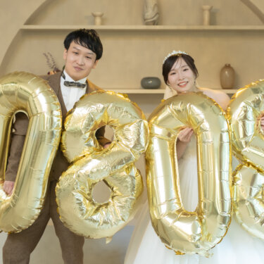 金色の数字バルーンで日付をかたどり、後ろから顔を出す新郎新婦。記念日を残せる小物使いのスタジオ前撮り