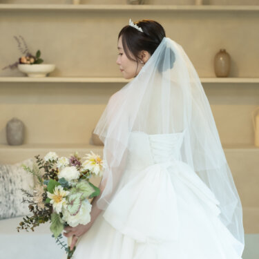 棚にオブジェが並ぶベージュのセットで、ブーケを手に振り返る花嫁の後ろ姿。ベールと編み上げのウェディングドレスで撮る前撮り