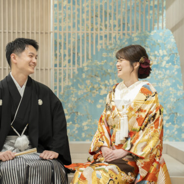 水色の花柄の壁を背にベンチに座って笑い合う黒紋付袴の新郎とオレンジの色打掛の花嫁。和装スタジオ前撮りの座りポーズ