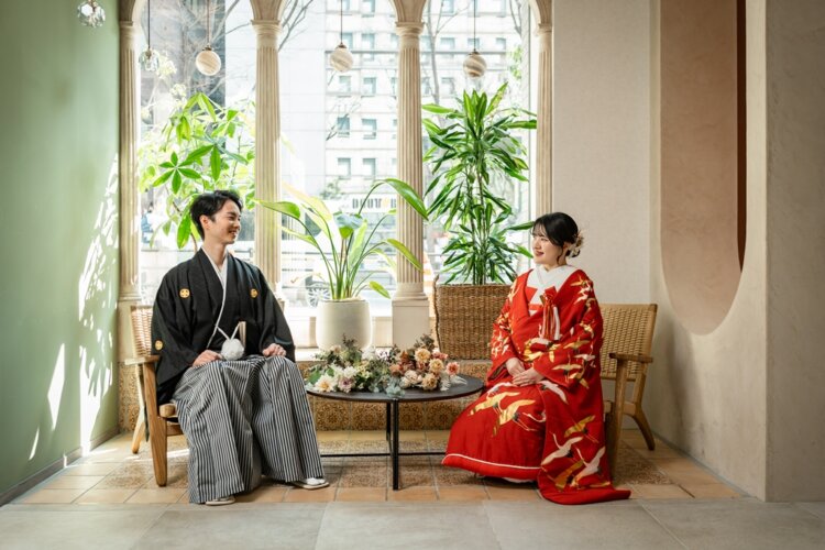 黒紋付の新郎と鶴の色打掛の花嫁が椅子に腰かけて見つめ合う横浜店のスタジオ。柱とグリーンに囲まれたフォトウェディング
