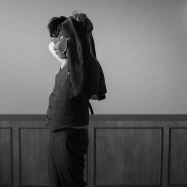 燭台の灯る壁の前で、ジャケットを肩にかけてポーズをとる新郎のモノクロ写真。ツイードのベストが映えるスタジオフォト