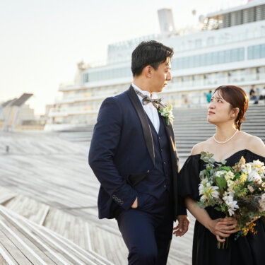 客船と大さん橋のデッキを背に、ブーケを抱えて新郎を見上げる黒いドレスの花嫁。見つめ合う自然なポーズのロケーションフォト