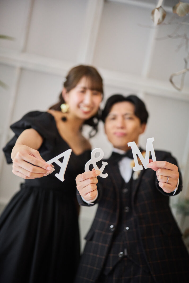 手前に『A&M』のイニシャルオブジェを掲げて笑うチェック柄のスーツの新郎と黒いカラードレスの花嫁。横浜のスタジオで撮る洋装前撮りのイニシャルのアイテムカット