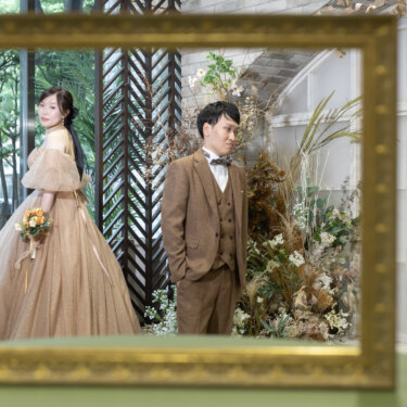手前の金の額縁越しに、窓辺に立つカラードレスの花嫁と新郎を写した構図。額縁フレームを生かしたおしゃれな前撮り