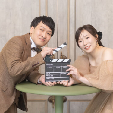 丸テーブルにもたれ、カチンコを持ってカメラを見る新郎新婦。ツイードのスーツとベージュのドレスで撮る遊び心あるスタジオフォト