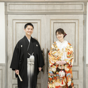白い扉の前で並んで立つ黒紋付袴の新郎とオレンジの色打掛の花嫁。スタジオで撮る和装前撮りの定番ポーズ