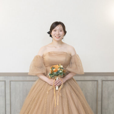 腰壁のあるスタジオの壁の前で、オレンジのブーケを持って立つベージュのカラードレスの花嫁。袖がふんわりしたチュールドレスの前撮り