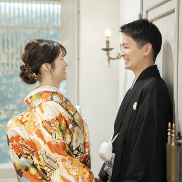 水色の花柄の壁を背に手をつないで見つめ合う色打掛の花嫁と黒紋付袴の新郎。和装スタジオフォトのやわらかな一枚